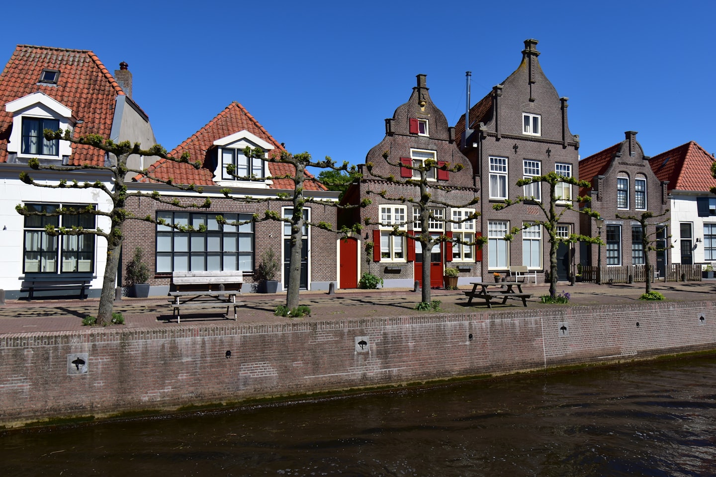 Gable-roofed houses line a canal with trees and picnic tables.