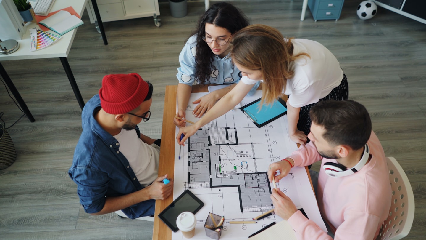 Vier ontwerpers werken samen aan een bouwtekening op tafel.